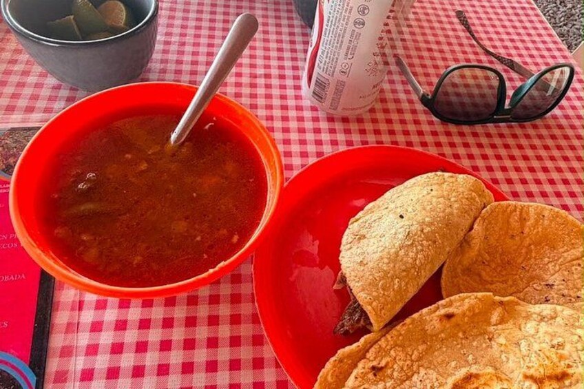 A red bowl of soup with a spoon, a red plate with two tortillas, a checkered tablecloth, and a pair of glasses. 3