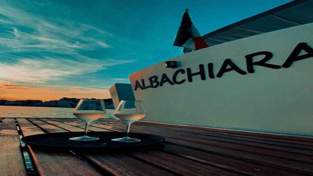 A boat with the name 'ALBACHIARA' on its side, two glasses of white wine on a tray, and a wooden deck. 2