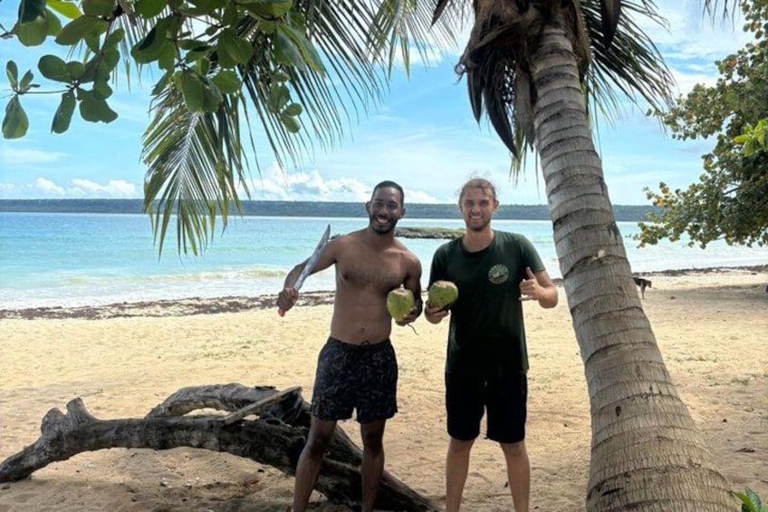 Two people on a beach holding coconuts, palm trees, and a large tree trunk. 4