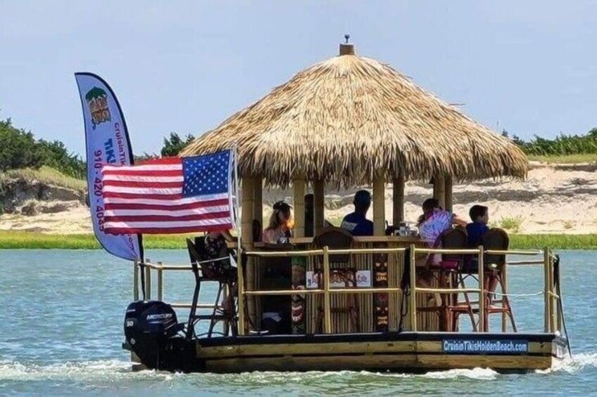 A boat with a thatched roof and an American flag, passengers, and a beach in the background. 1