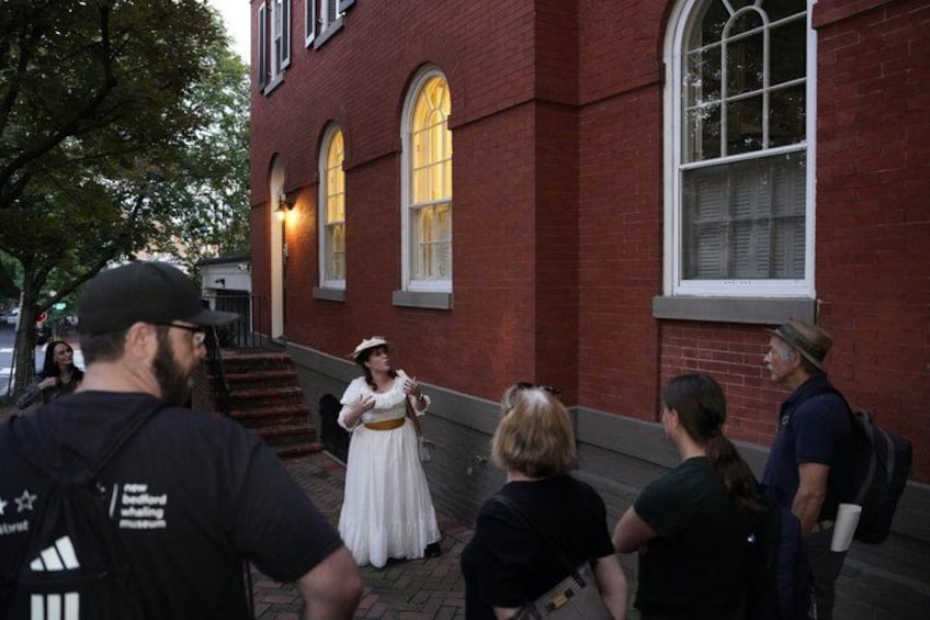 A person in historical costume standing on a sidewalk in front of a red brick building with arched windows. 4