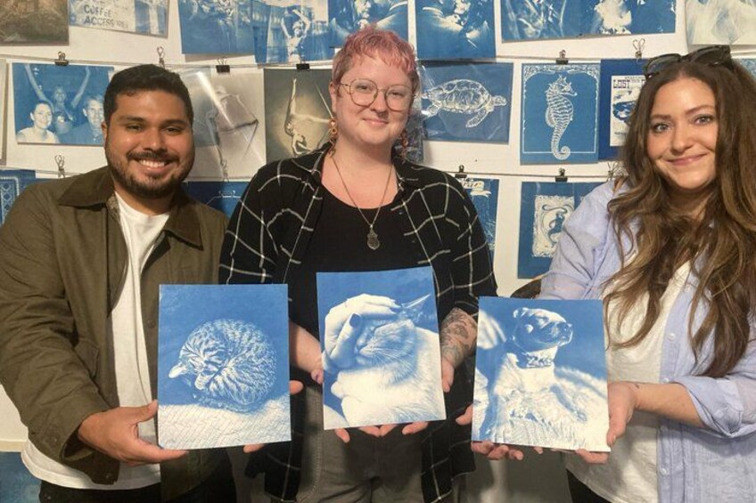 Three people holding framed artwork against a wall with blue and white prints. 1