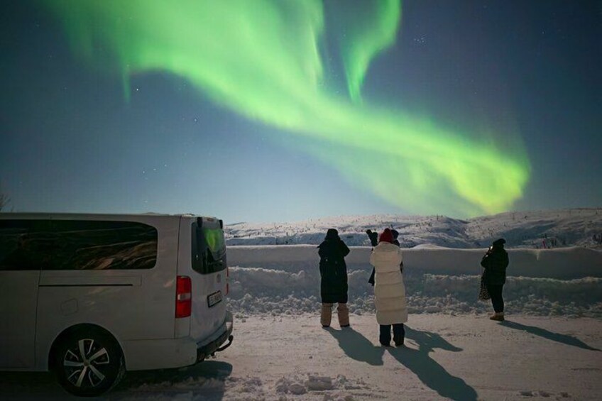 A white van parked on a snowy landscape with people observing the northern lights. 1