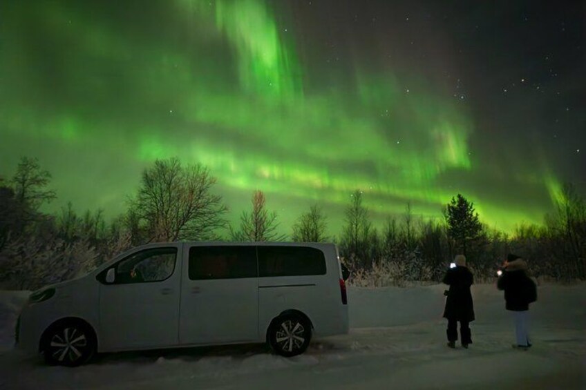 A white van parked on a snowy road with people observing the northern lights. 2