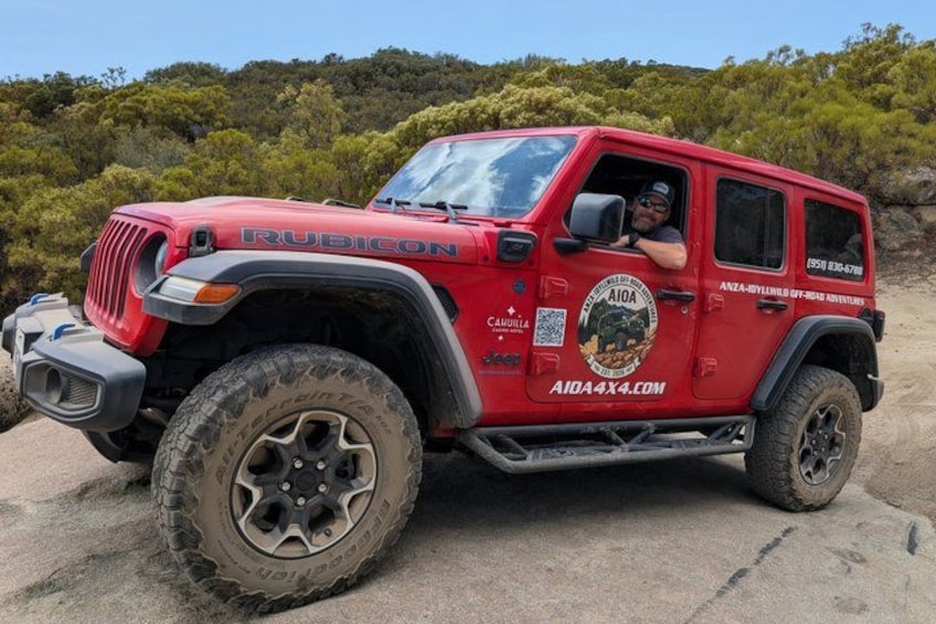A red Rubicon Jeep with stickers and decals, parked on a dirt surface with greenery in the background. 2