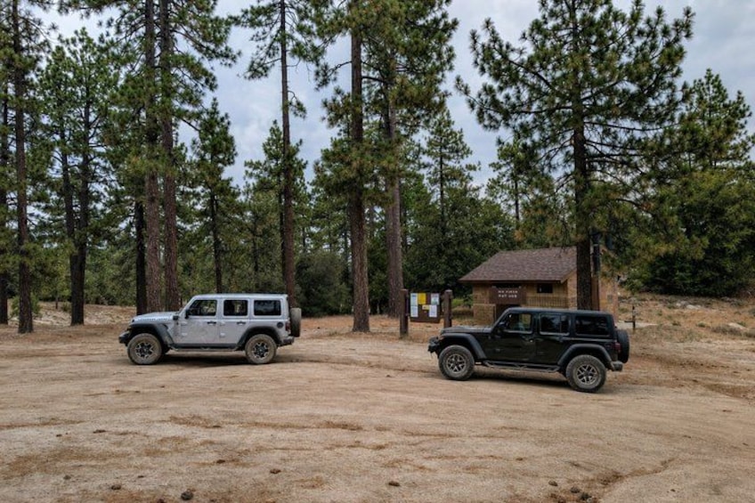 Two off-road vehicles parked in a forested area with a small cabin in the background. 1
