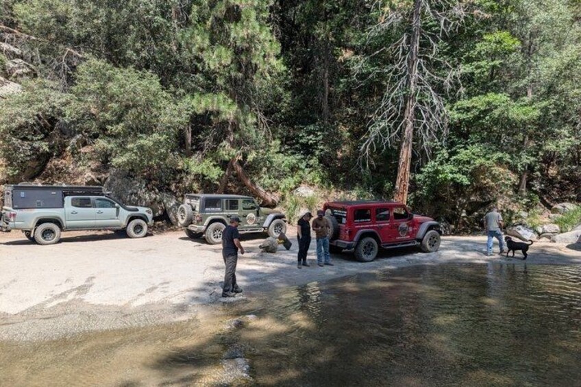 A group of people near a river with two off-road vehicles parked on the shore. 1