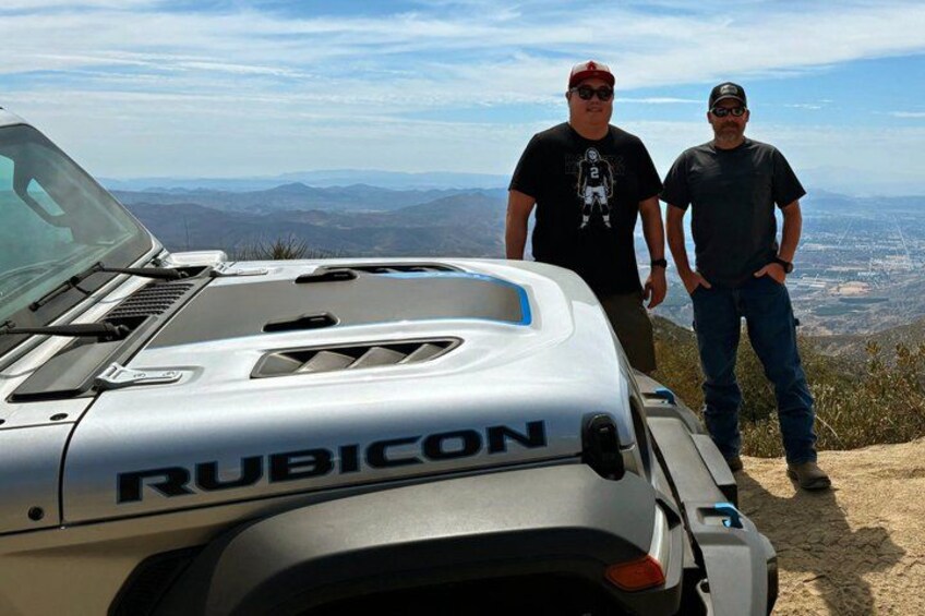 A Rubicon vehicle with two people standing beside it on a mountain overlooking a valley. 5