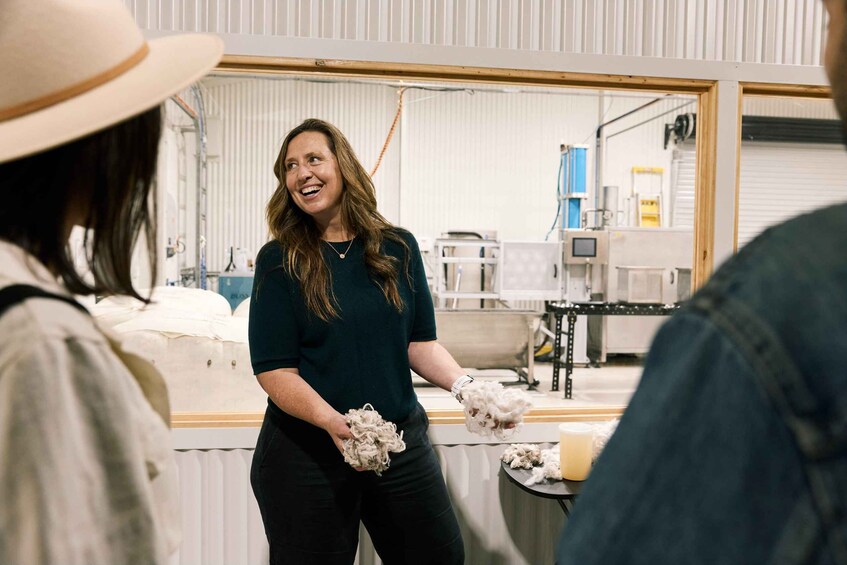 A person holding a pile of white material, possibly wool, in a workshop with industrial equipment in the background. 1