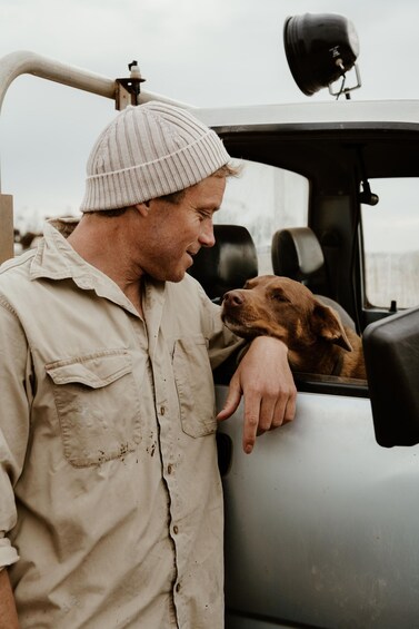 A man in a beige shirt and a beanie leaning on a vehicle with a brown dog resting on the side. 3