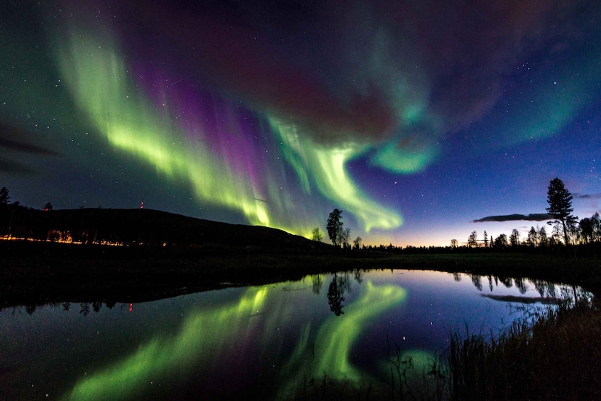 The Northern Lights over a calm lake with reflections, surrounded by trees and a distant hill. 1
