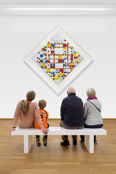 Eine vierköpfige Familie sitzt auf einer Bank und betrachtet ein farbenfrohes, geometrisches Kunstwerk an einer Wand. 4