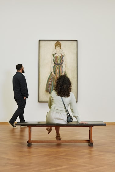 Eine Person sitzt an einem Tisch in einer Kunstgalerie und betrachtet ein Gemälde einer Frau in einem farbenfrohen Kleid. 3