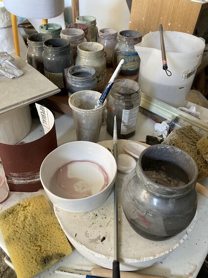 A cluttered workspace with pottery tools, jars, and a bowl of liquid. 2