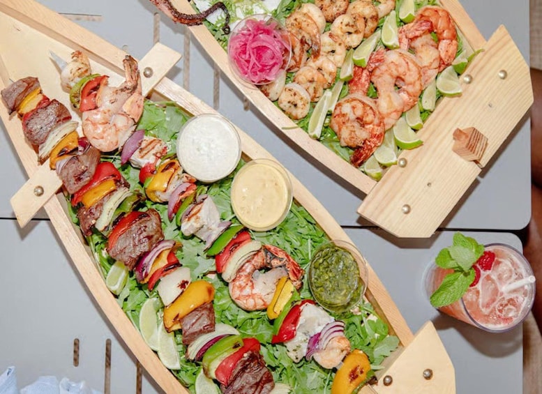A wooden serving tray with a variety of grilled meats, shrimp, and vegetables, accompanied by a drink and a small bowl of sauce. 5