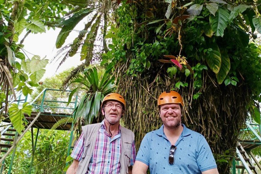 Two people wearing helmets, standing in a lush, green environment with dense foliage and a metal staircase. 5