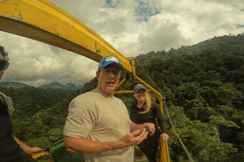A person in a beige shirt and blue cap standing next to a yellow excavator in a forested area with a cloudy sky. 1