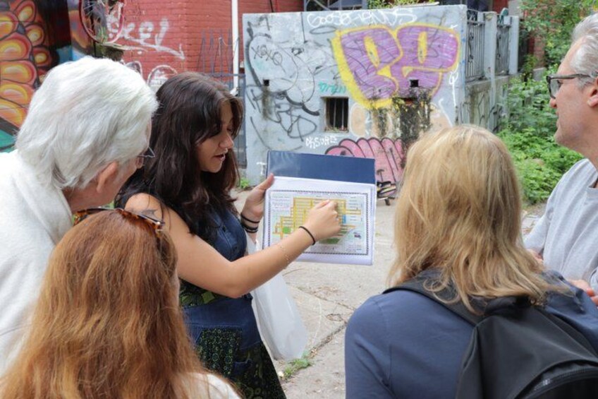 A group of people gathered around a woman holding a map, standing in an alley with graffiti on the walls. 3