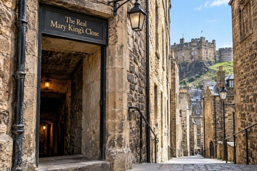 A narrow street with stone buildings, a sign reading 'The Real Mary King's Close,' and a castle in the background. 4