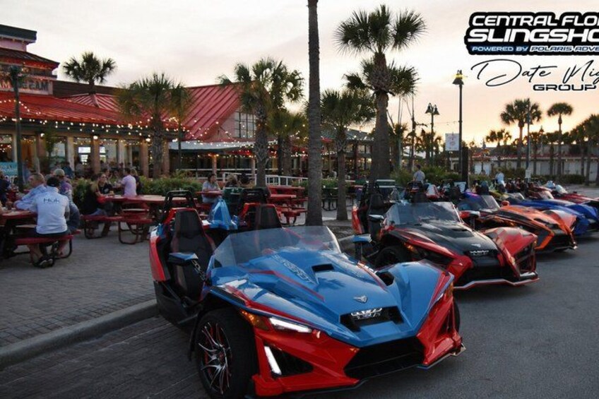 A row of sports cars parked outdoors, palm trees, and a building with a red roof in the background. 1