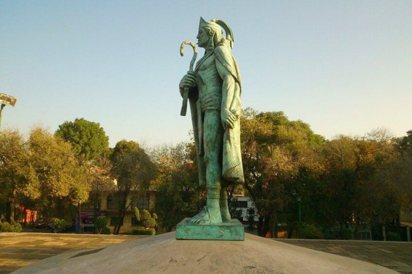 A statue of a person in traditional attire holding a staff, set in a park with trees and a clear sky. 3