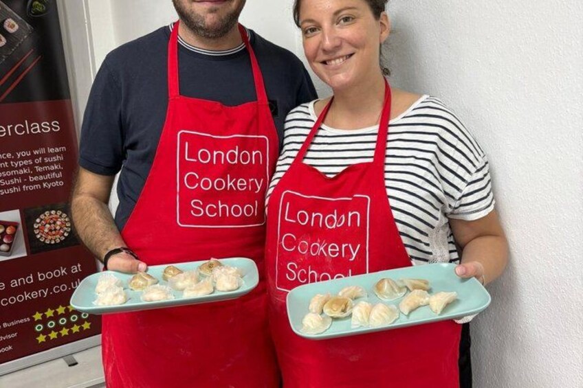 Two people holding plates of food, wearing red aprons with 'London Cookery School' text, in front of a sign with the same text. 5