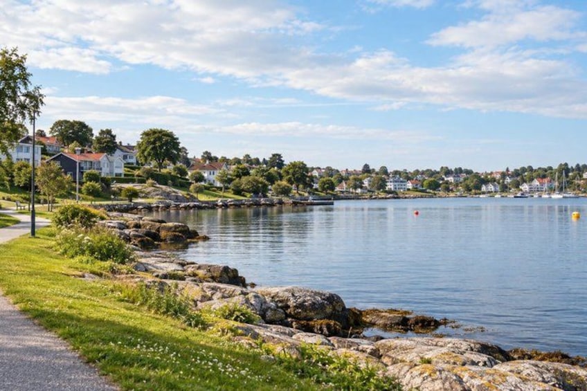 A calm lake with a rocky shoreline, a grassy path, and houses in the background. 1