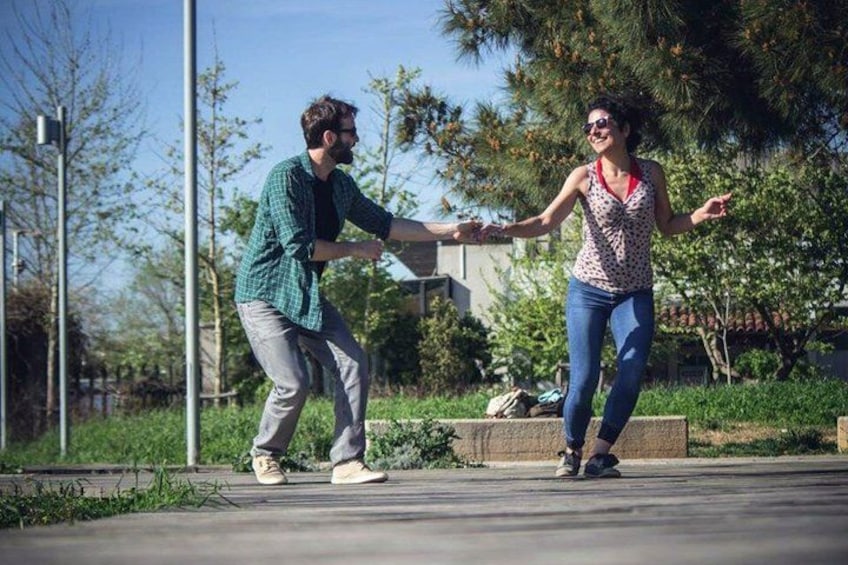 Two people dancing on a concrete path in a park with trees and a building in the background. 1