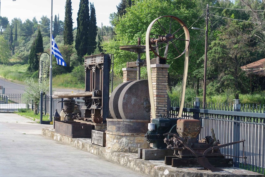 An old oil press with a large wheel and mechanical parts, surrounded by a stone wall and a metal fence. 1