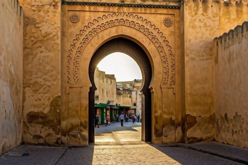 An arched doorway with intricate carvings, leading to a cobblestone street with buildings and people in the background. 4