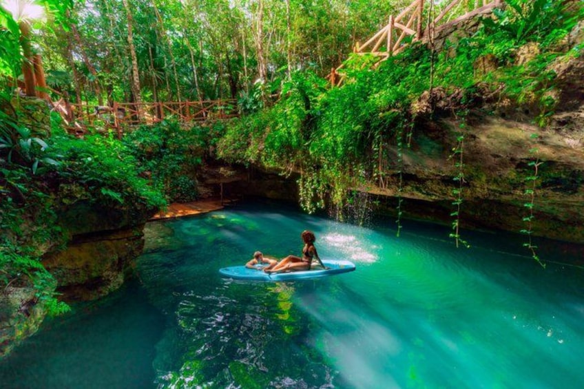 A person floating on a raft in a clear blue river surrounded by lush greenery and a wooden bridge. 1