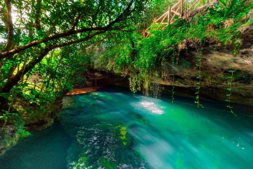 A natural cave with clear water, surrounded by lush greenery and a wooden bridge. 2