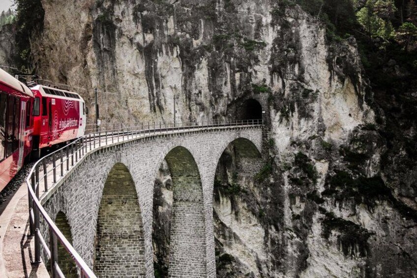 A red train on a stone bridge with multiple arches, surrounded by rocky cliffs. 5