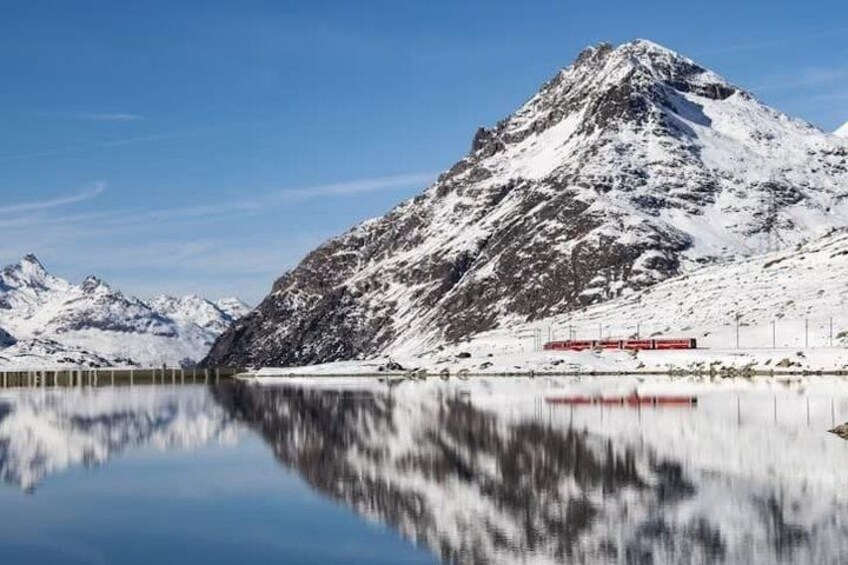 A snow-covered mountain, a calm lake, and a red train. 1