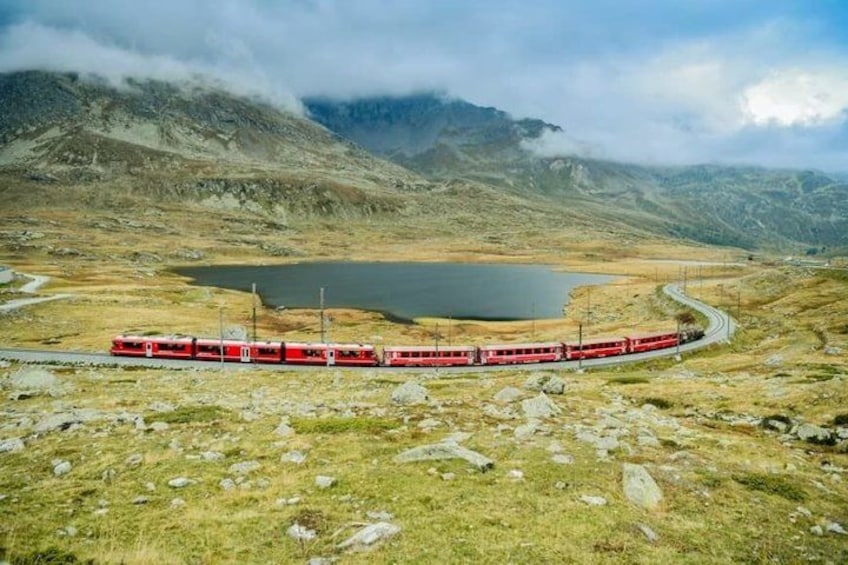 A red train on a mountainous track with a lake and clouds in the background. 4
