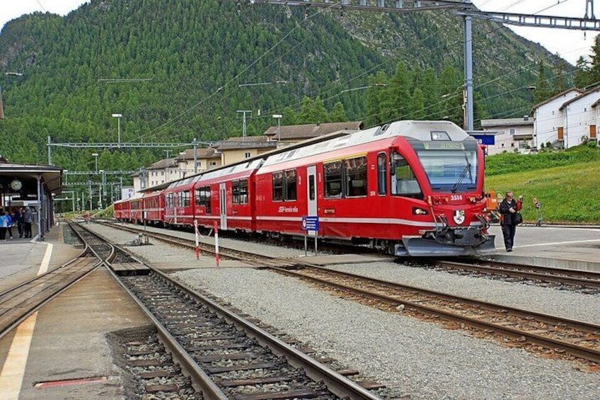 A red train at a station with tracks, a platform, and a mountainous backdrop. 2