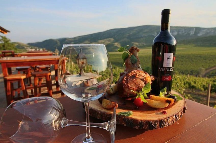 A wine glass, a bottle of Merlot, and a roasted chicken on a wooden board with a scenic vineyard in the background. 1