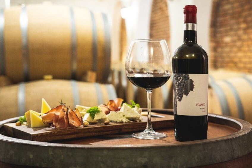 A wine bottle, a glass of red wine, and a wooden tray with food items on a round table in a winery setting. 3