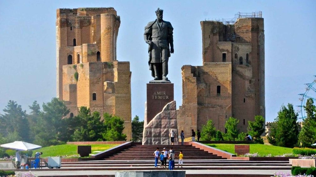 Une statue d’un homme en costume, portant une couronne, est dressée sur un piédestal portant l’inscription « AMIR TEMUR », devant deux grandes tours en briques. 1
