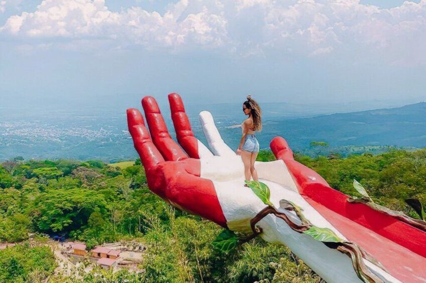 A person standing on a large hand-shaped structure with red and white colors, overlooking a landscape with greenery and distant hills. 1