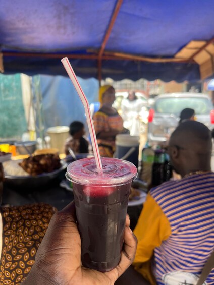 A hand holding a plastic cup with a straw, filled with a dark beverage, in front of a market scene. 4