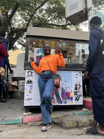 A person in an orange sweater and jeans posing in front of a kiosk with posters. 1