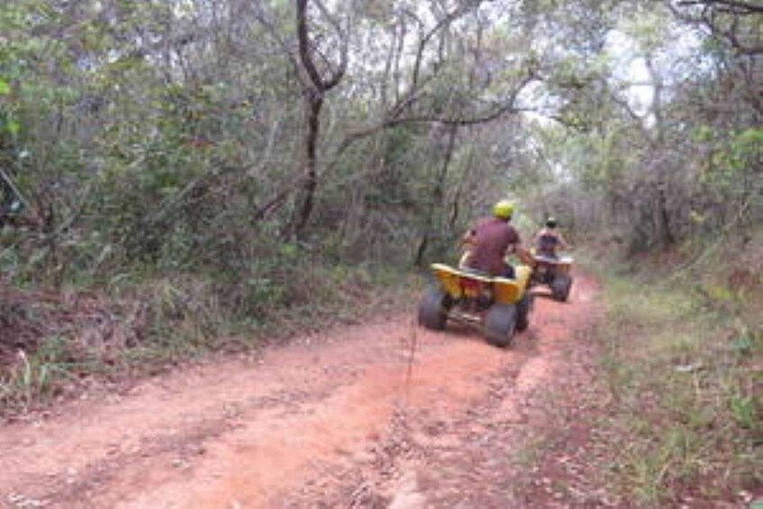 Two people riding off-road vehicles on a dirt path through a wooded area. 3