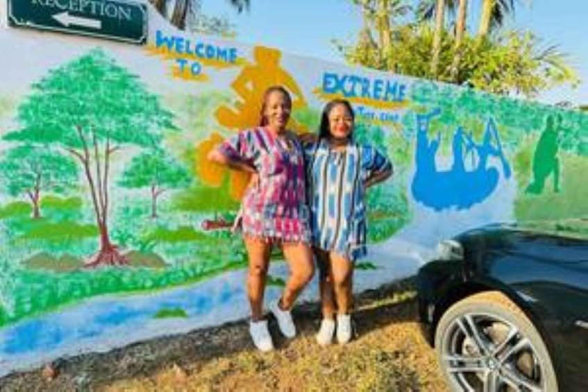 Two people standing in front of a colorful mural with trees and a car. 2