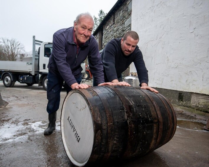 Two men pushing a large wooden barrel on a wet surface with a truck and a building in the background. 5