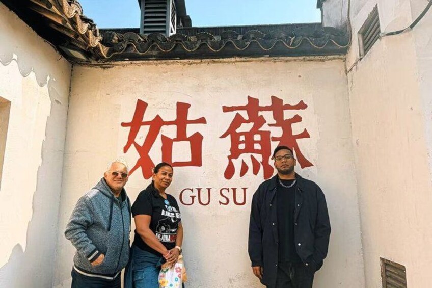 Three people standing in front of a wall with large red Chinese characters and the word 'GU SU' in smaller text. 1