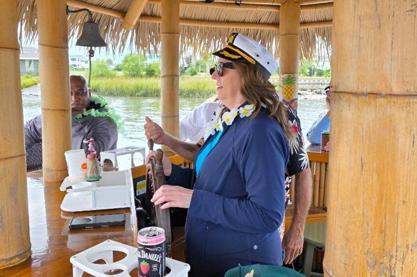A person in a blue shirt and white hat standing at a wooden table with a drink and a small figurine. 2