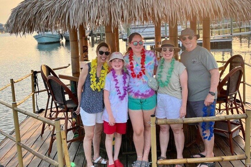 A group of people standing on a wooden dock with a thatched roof, overlooking calm water. 4