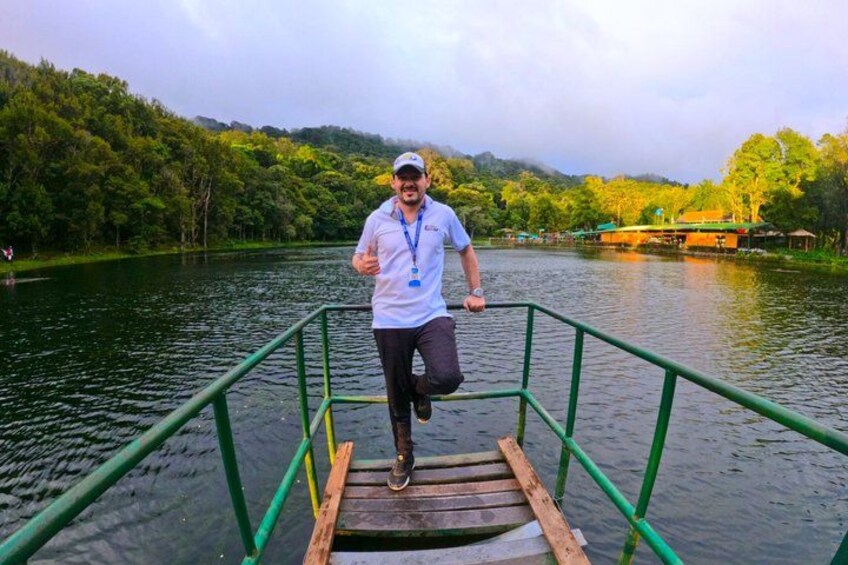 A person walking on a wooden platform with green railings over water, surrounded by trees and a building in the background. 4