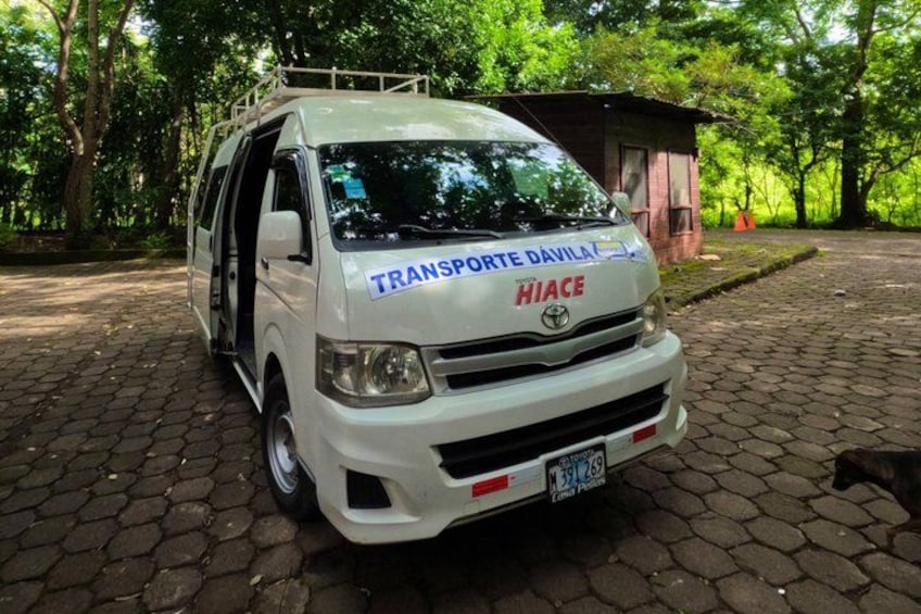 A white van with 'TRANSPORTE DAVILAT' and 'HIACE' written on it, parked on a cobblestone path with trees in the background. 2